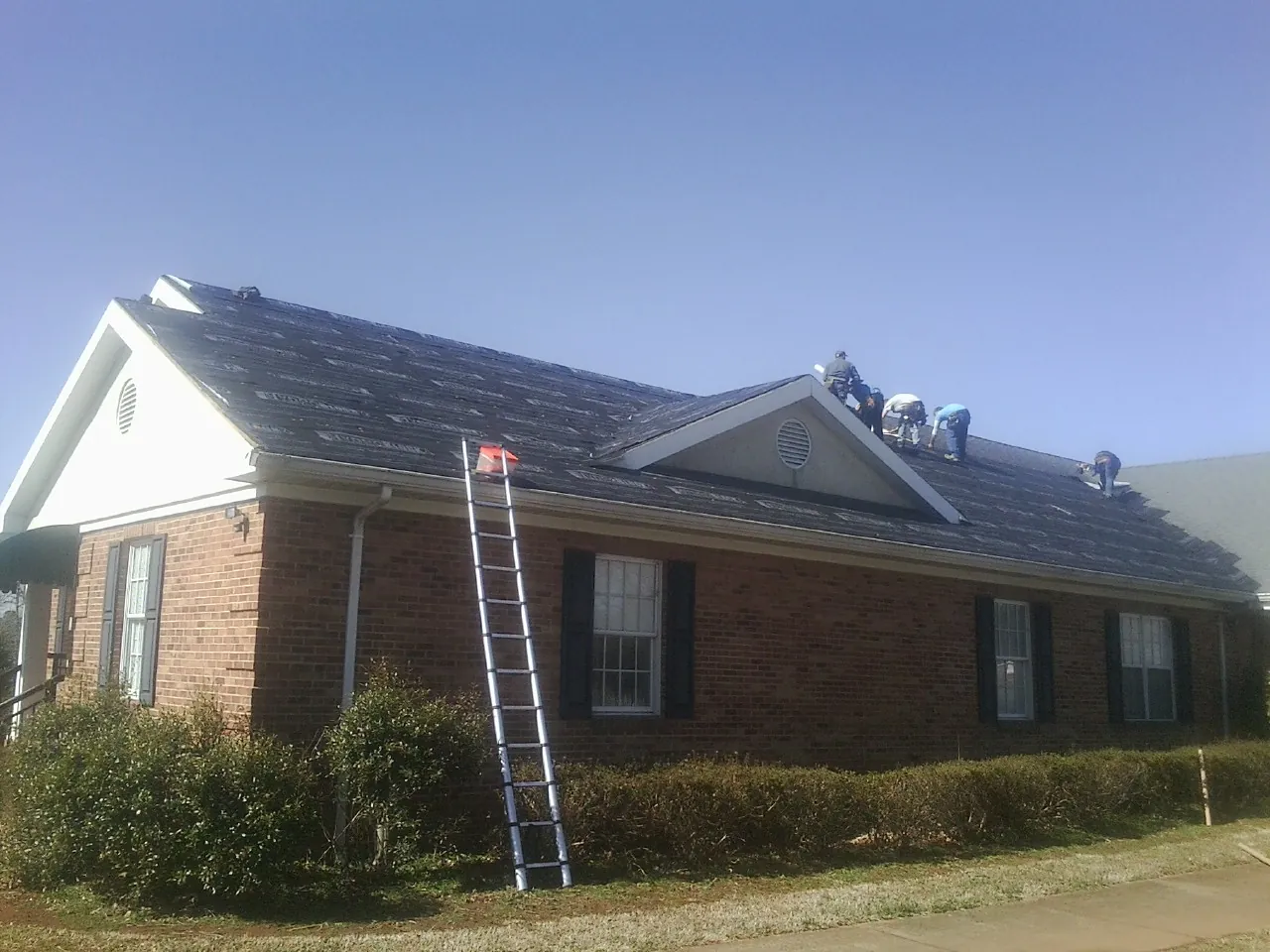 Crew tearing shingles off a brick ranch roof with a ladder set — Metal Roof Repair work by our crew in Horace